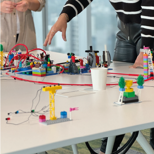 Personas construyendo modelos simbólicos de cambio y colaboración en una sesión de LEGO Serious Play, con vistas a una ciudad desde oficinas corporativas